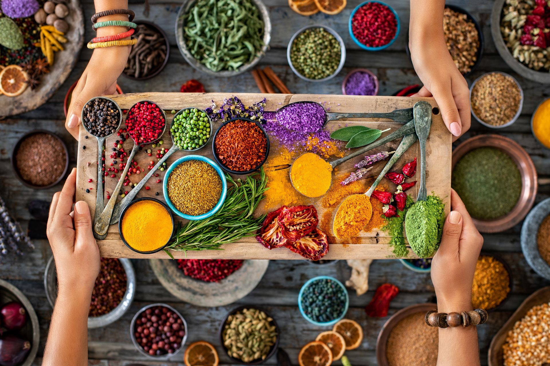 Origins matter: An image of a pallet of spices including curry spices, pepper, macha, sun dried tomatoes, on a wooden tray being held by two people